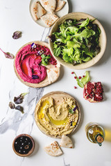 Variety of homemade traditional and beetroot spread hummus with pine nuts, olive oil, pomegranate served on ceramic plates with pita bread and green salad on white marble background. Flat lay, space.