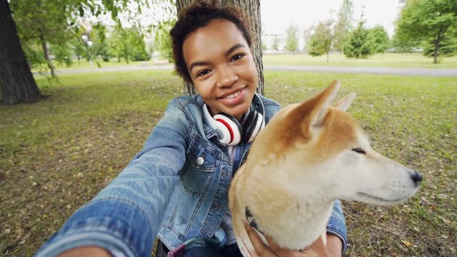 Point of view shot of pretty African American girl taking selfie with cute puppy in city park holding camera, smiling and posing. Modern technology and animals concept.