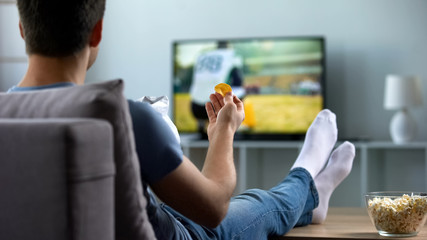Man watching american football eating snacks on couch home, weekend leisure - Powered by Adobe