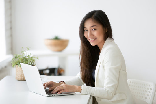 Portrait of beautiful Asian female office worker looking at camera smiling while working at laptop, young businesswoman posing for company catalogue near desk. Concept of leadership, ethnicity