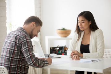 Asian female employer watching male job candidate signing work contract after successful interview in office, man closing partnership deal, putting signature on document. Hiring, recruitment concept