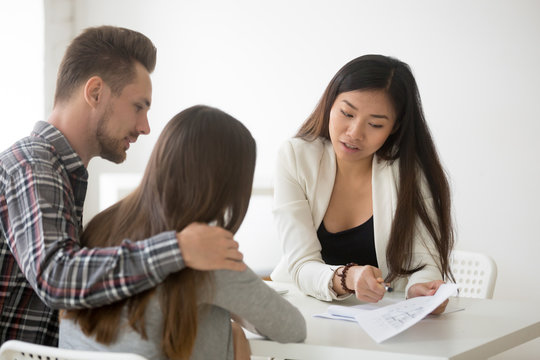 Female Asian Broker Show House Plan To Happy Millennial Couple Deciding To Buy First Home Together, Spouses Visiting Realtor To Purchase Property, Man And Woman Listening To Real Estate Agent Talking