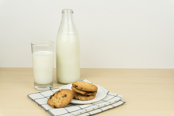 a glass of milk with milk bottle and the chocolate chip cookies in a white round dish on the wooden table.