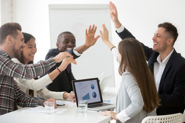 Excited diverse work team giving high five at board meeting, celebrating shared goal achievement,...
