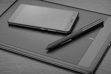 Smartphone and graphic tablet (also known as a digital drawing tablet) with a stylus on a wooden table. Details of workplace of an digital graphics art designer
