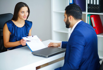 Business. Business Woman and Business Man Talks at the Light Office Sitting at the Table. Woman Dressed in Blue Blouse and Beige Skirt. Man Dressed in Blue Costume and White Shirt. High Resolution