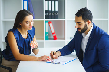 Business. Business Woman and Business Man Talks at the Light Office Sitting at the Table. Woman Dressed in Blue Blouse and Beige Skirt. Man Dressed in Blue Costume and White Shirt. High Resolution