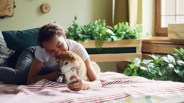 Smiling mixed race teenager is taking selfie with puppy then talking to it and showing it photos on smartphone screen lying on bed at home. Technology and pets concept.