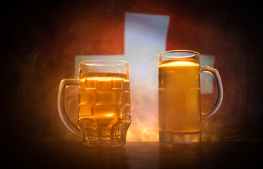 Creative concept. Beer glasses on table at dark toned foggy background with blurred view of flag of Switzerland. Support your country with beer concept.