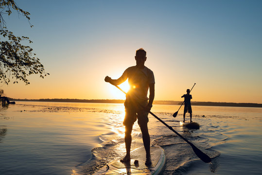 Men, friends sail on a SUP boards in a rays of rising sun