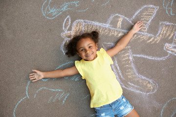 Little African-American child lying near chalk drawing of airplane on asphalt, top view
