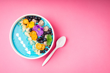 Blue smoothie bowl with fruits, berries, nuts and flowers on pink background. Tropical healthy smoothie dessert. Healthy eating, vegetarian, diet concept. Top view