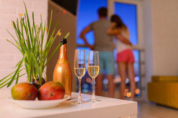 Bottle and two glasses of champagne on coffee table in living room, couple in love embraced at background and looking thru window to night city.