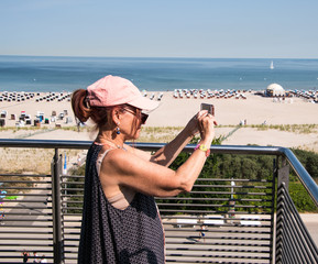 Woman baby boomer wearing pink baseball cap on terrace taking picture of the beach and ocean with her call phone