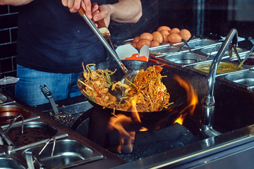 Cooking process in an Asian restaurant. Cook is fry vegetables with spices and sauce in a wok on a...