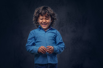 Laughing cute schoolboy with brown curly hair dressed in a blue shirt, posing at a studio. Isolated on a dark textured background.