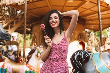 Beautiful lady with dark curly hair in dress standing with lolly pop candy in hand while happily looking in camera with carousel on background © Anton