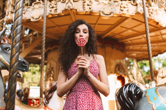 Beautiful lady with dark curly hair in dress standing and covering her mouth with candy while dreamily looking in camera with carousel on background