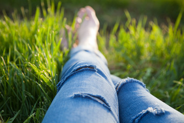 legs of hipster woman wearing torn jeans lying in green grass with smartphone and headsets enjoying sunset - summertime relaxation