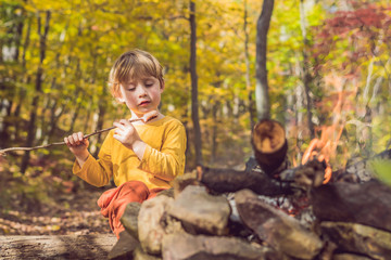 The boy is sitting at the camp fire and eating a fried sausage