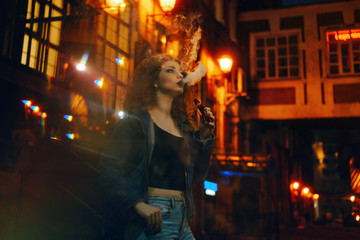Fototapeta premium Young attractive red-haired girl smokes electronic cigarette. She walks through small streets of city. Evening city lights blink.