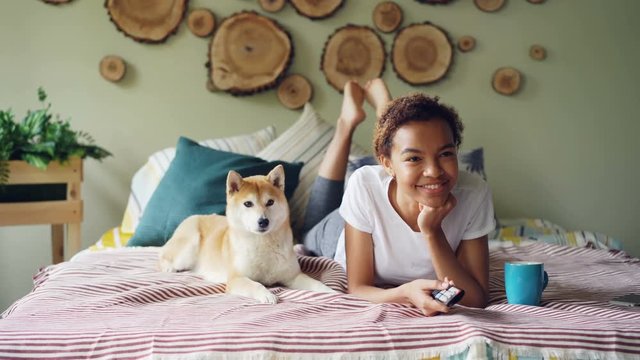 Cheerful mixed race student is watching TV holding remote and pressing buttons choosing television channels while her dog is moving on bed at home.