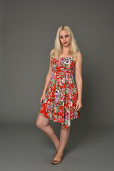 full length portrait of blonde girl wearing red floral dress, standing pose on grey studio background.