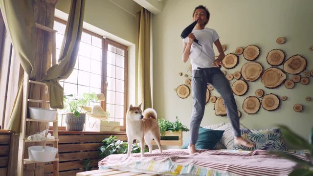 Funny African American girl is having fun listening to music, jumping and dancing on bed, singing in hair dryer with beautiful shiba inu dog walking on bed.