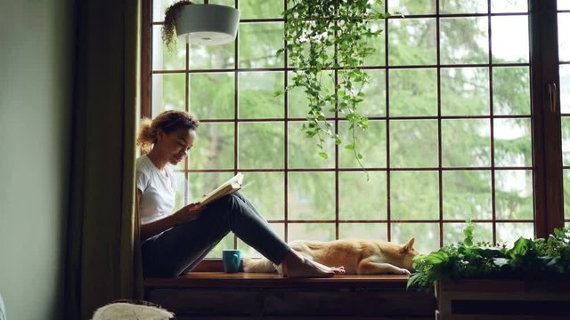 Attractive girl in casual clothes is reading book sitting on window sill barefoot near adorable pet dog lying near her owner. Home, hobby and animals concept.