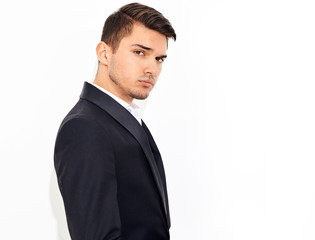 portrait of handsome fashion stylish hipster lumbersexual businessman model dressed in elegant black classic suit posing near white wall in studio. Metrosexual