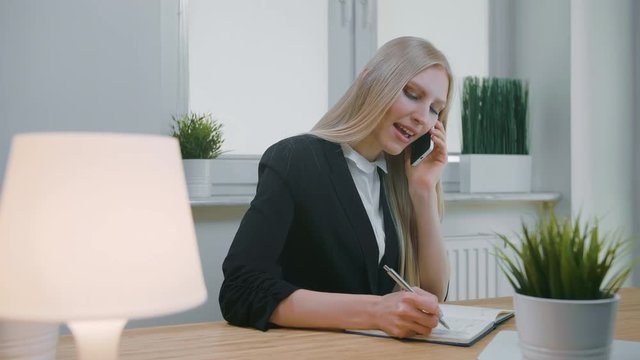 Business woman talking on smartphone in office. Elegant young blond female in office suit sitting at workplace and negotiating via mobile phone in hand writing down necessary information into notebook