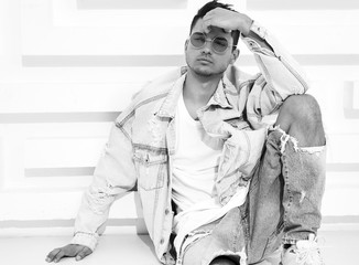 Portrait of handsome young model man dressed in jeans clothes sitting near white textured wall