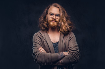 Close-up portrait of a redhead hipster male with long luxuriant hair and full beard dressed in casual clothes standing in a studio, looking away. Isolated on the dark background.
