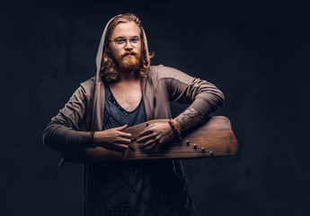 Redhead hipster male with long luxuriant hair and full beard dressed in a hoodie and t-shirt playing on a Russian traditional musical instrument - gusli. Isolated on a dark background.