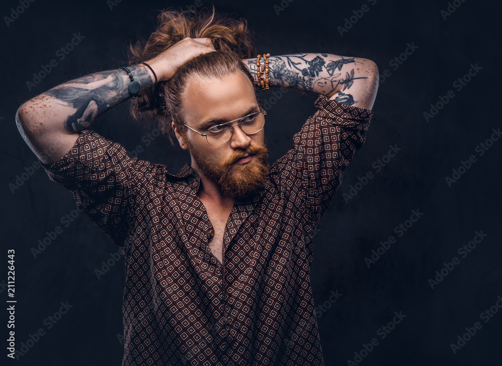 Wall mural Tattoed redhead man hipster corrects his lush hair dressed in a brown shirt, standing at a studio. Isolated on a dark background.