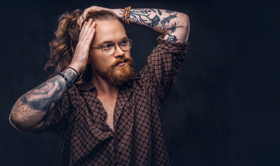 Tattoed redhead man hipster corrects his lush hair dressed in a brown shirt, standing at a studio. Isolated on a dark background.