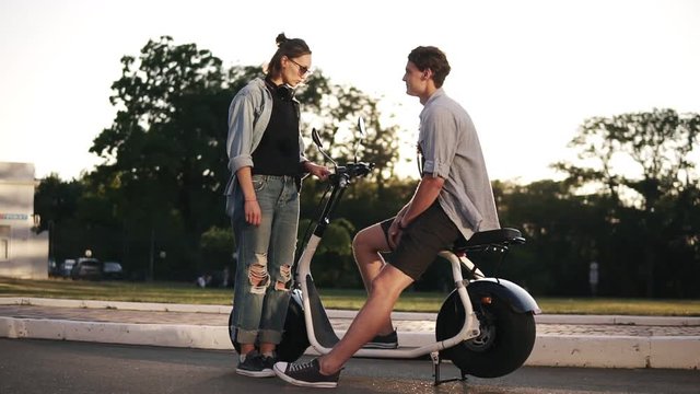 Young male and female are have time together outdoors. Boy's sitting on an electro mini bike and smiling. The girl in sunglasses stands near and talks. Sun shines on the background