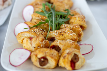 Snacks from puff pastry with sausage in the middle, sprinkled with sesame, decorated with arugula and radish