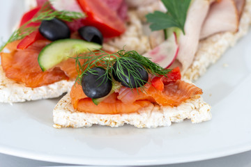 Sandwich with smoked salmon served on a rice waffle decorated with olive, cucumber, paprika and a sprig of fennel on a white plate closeup