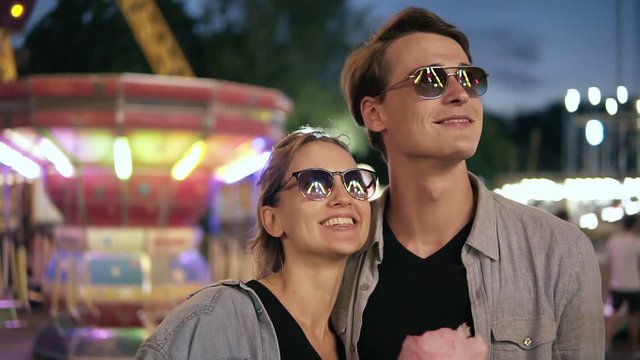 Portrait of a young, cheerful hipster couple in sunglasses are looking at attractions light. Happy, smiling young people at funfair at night