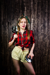 Young funny housewife in checkered shirt and yellow shorts pin up style with saucepan on wooden background.