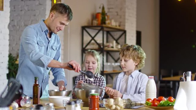 Smiling father adding flour to metal container when cooking with two children, who are holding sieves