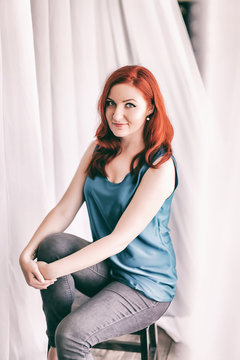 Image of redhead young happy lady in casual clothes sitting on chair on blurred white background looking camera