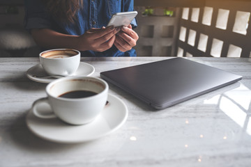 Closeup image of a woman holding and using smartphone with coffee cups and laptop on table in cafe