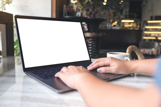 Mockup image of a woman using and typing on laptop with blank white screen and coffee cup on table in modern cafe