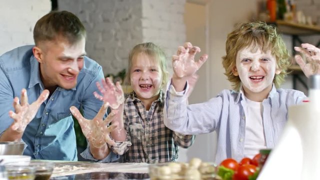 Medium shot of happy father and two children playing with flour in kitchen and making crazy faces at camera