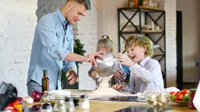 Young Caucasian man in denim shirt scattering flour on kitchen worktop and letting two children play with it when baking food together