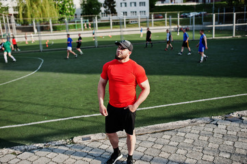 Fototapeta premium Young brutal bearded muscular man wear on red shirt, shorts and cap at stadium.
