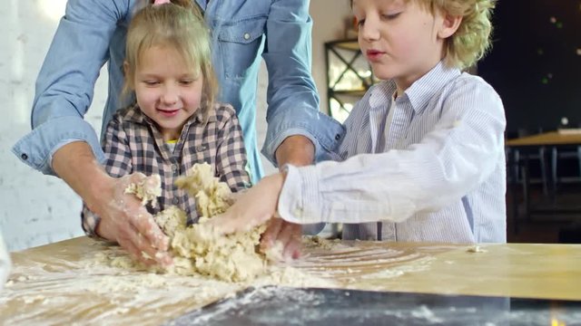 Handheld shot of two joyful children and their father mixing ingredients with hands when making dough for bakery together