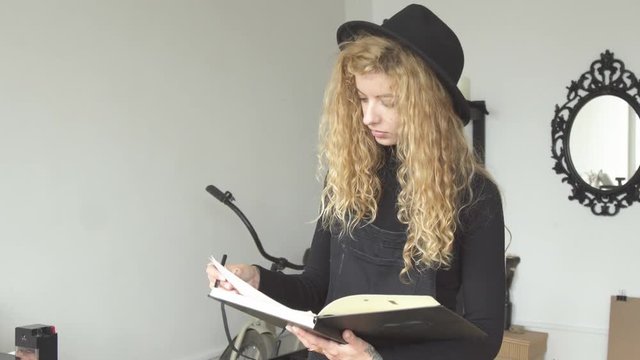 Young caucasian painter in cute black hat making a sketch in the sketch album, preparing for the next work of art as standing opposite the mirror in small art studio
